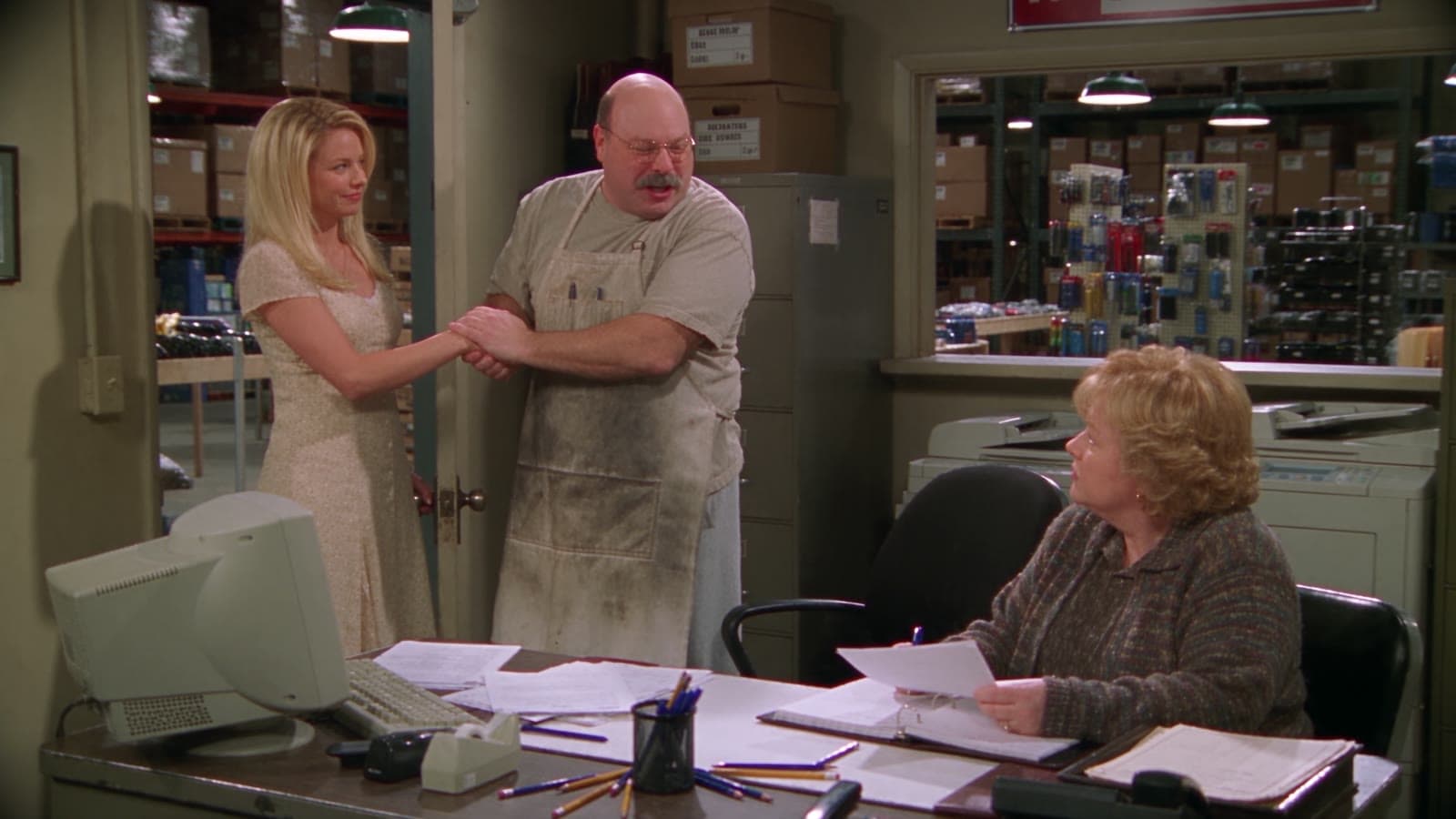 Gary schüttelt einer jungen Frau im Büro des Ladens die Hand und grinst, Linda sieht vom Schreibtisch aus zu