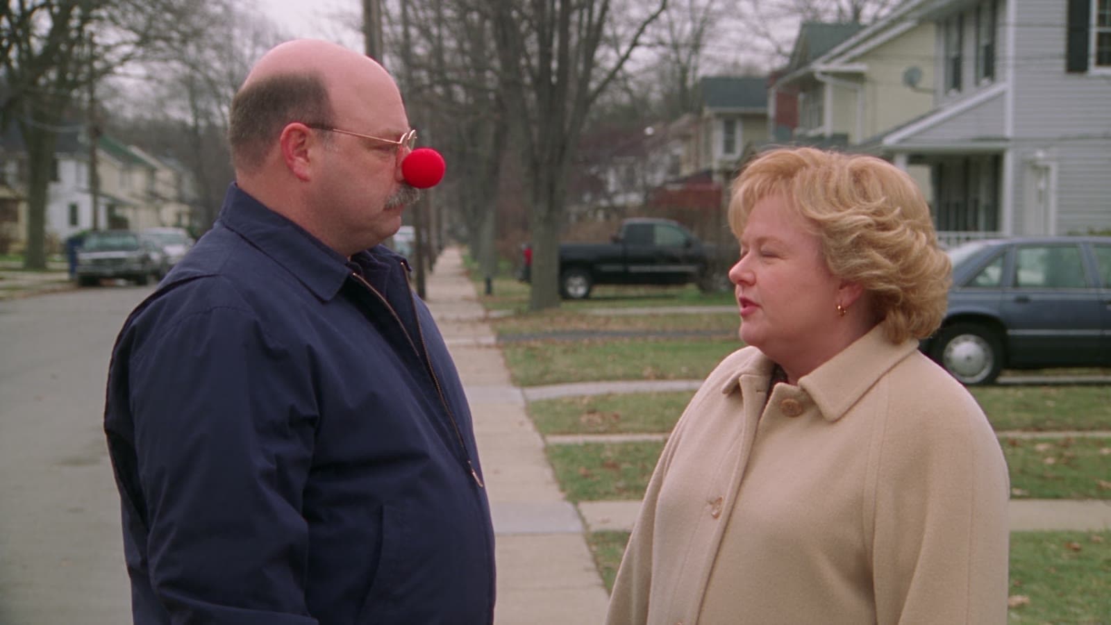 Gary und Linda stehen mit Jacken auf einem Gehweg, Gary trägt eine rote Clownsnase und schaut Linda ernst an