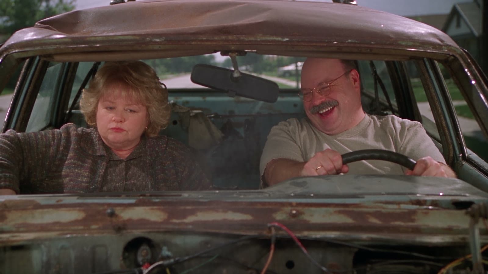 Gary und Linda in einem verrosteten Auto, er grinst sie von der Seite an