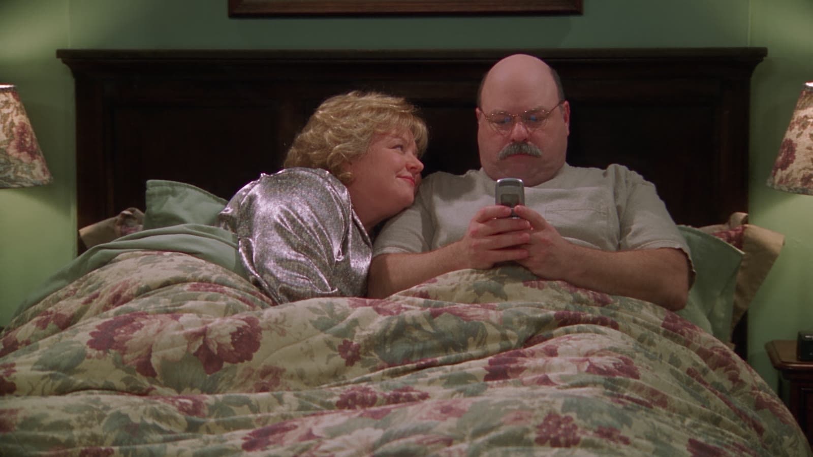 Linda schmiegt sich im Bett an Gary, der auf sein Handy starrt