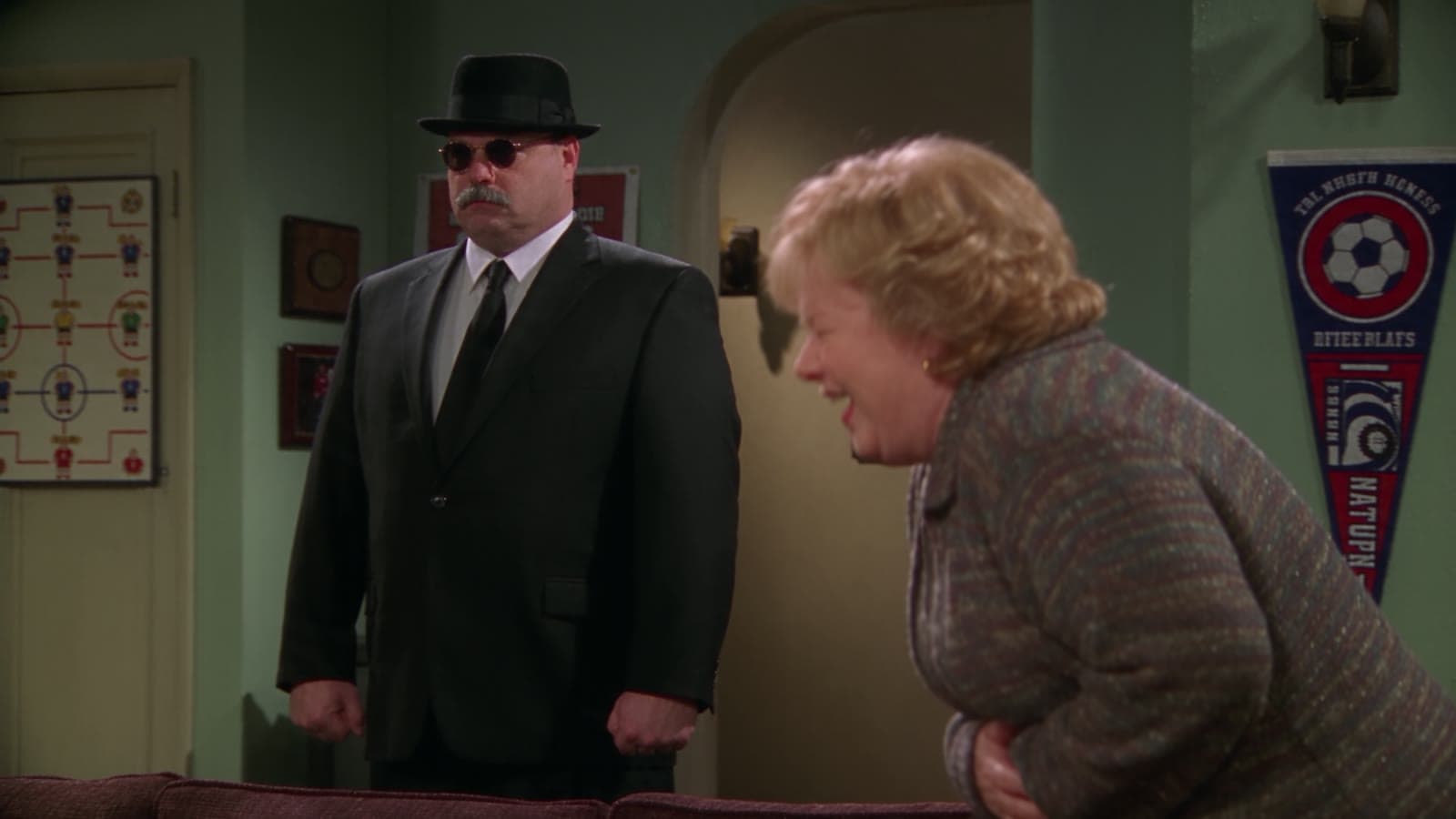 Gary steht im schwarzen Anzug mit Hut und Sonnenbrille im Wohnzimmer, Linda lacht