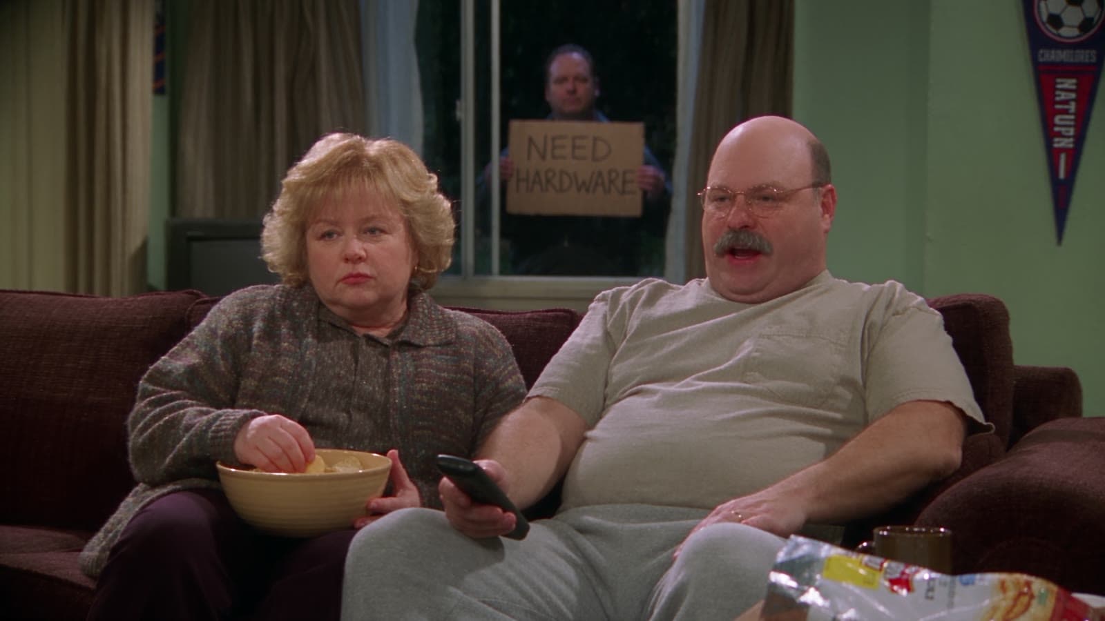 Gary und Linda auf der Couch, hinter ihnen steht nachts ein Mann am Fenster mit einem Pappschild