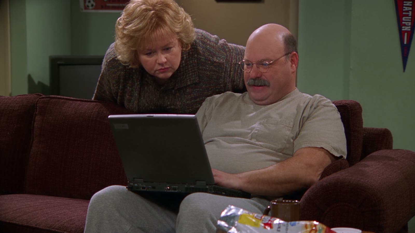 Gary sitzt mit Laptop auf der Couch, Linda schaut ihm über die Schulter auf den Bildschirm